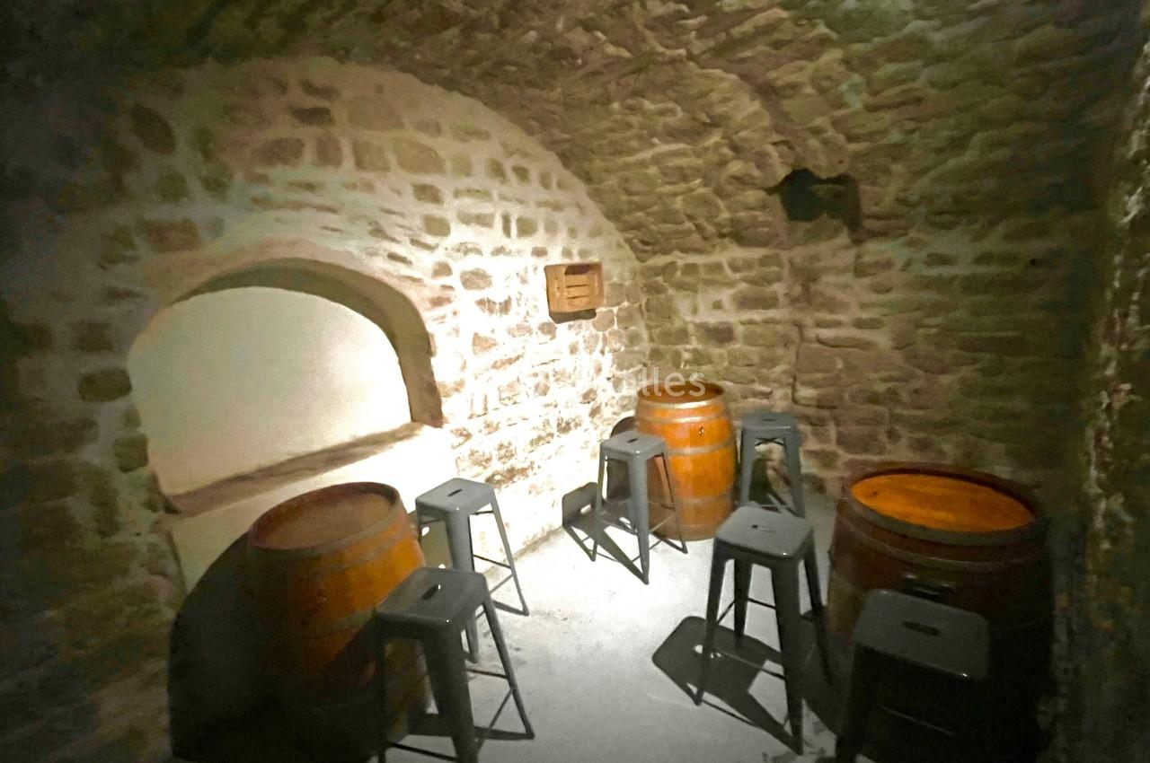 Cave voûtée en pierre avec des tonneaux en bois utilisés comme tables entourés de tabourets métalliques.