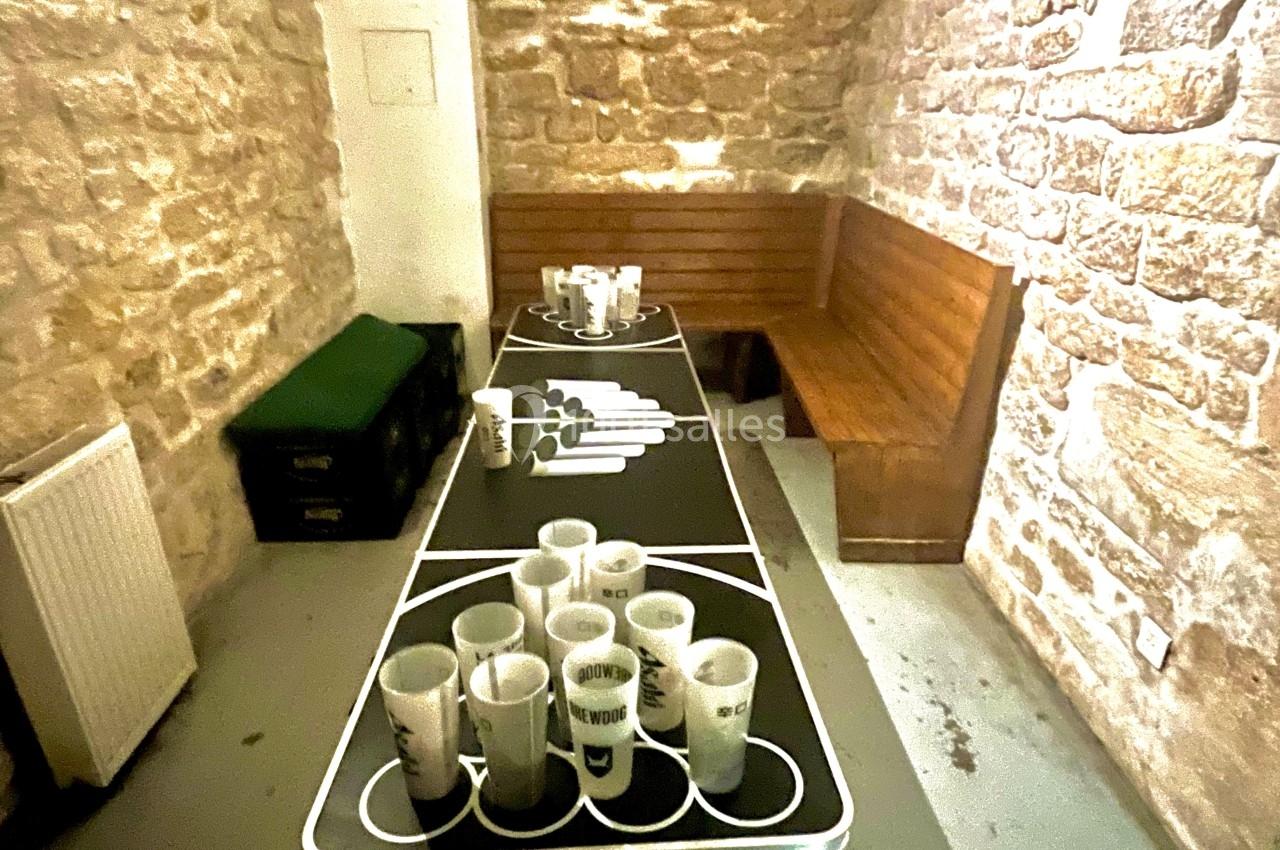 Table de beer-pong avec gobelets disposés, dans une pièce aux murs en pierre et bancs en bois.