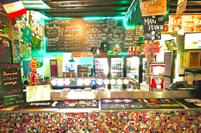 Intérieur d'un bar décoré de fanions, affiches et stickers, avec des tables en bois et des tabourets métalliques.