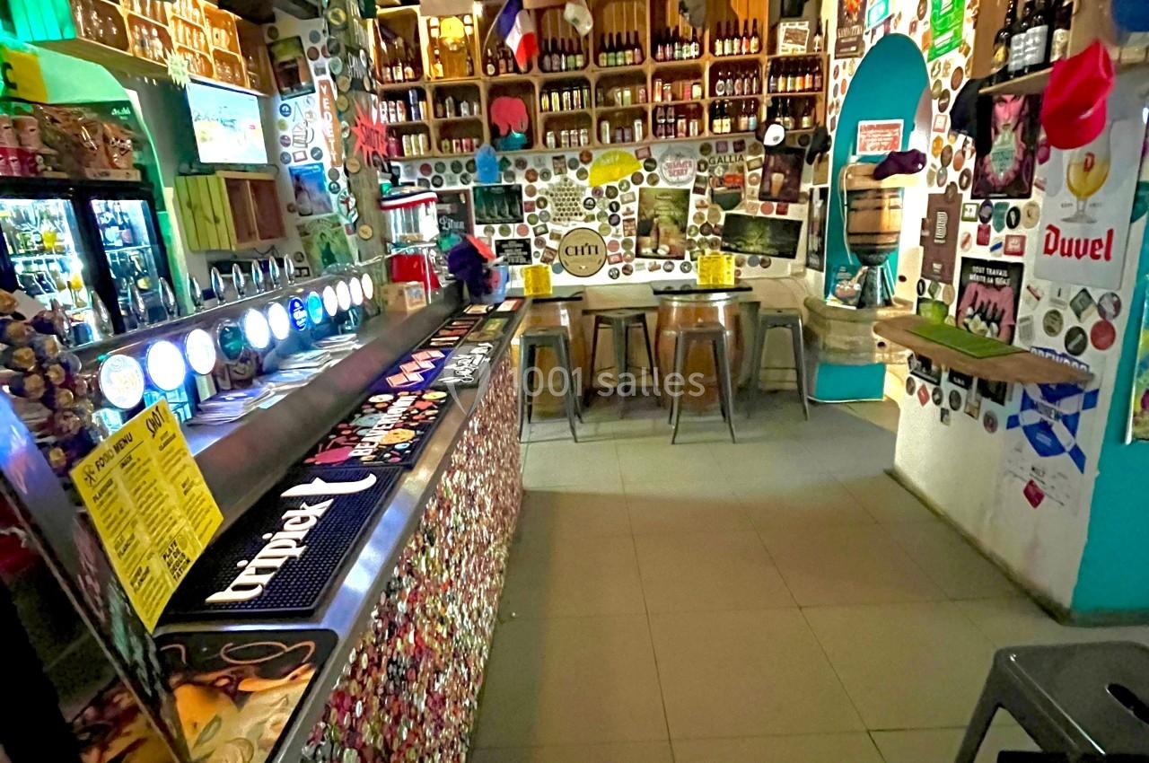 Intérieur d'un bar décoré avec des drapeaux, des bouteilles exposées et un long comptoir orné de capsules.