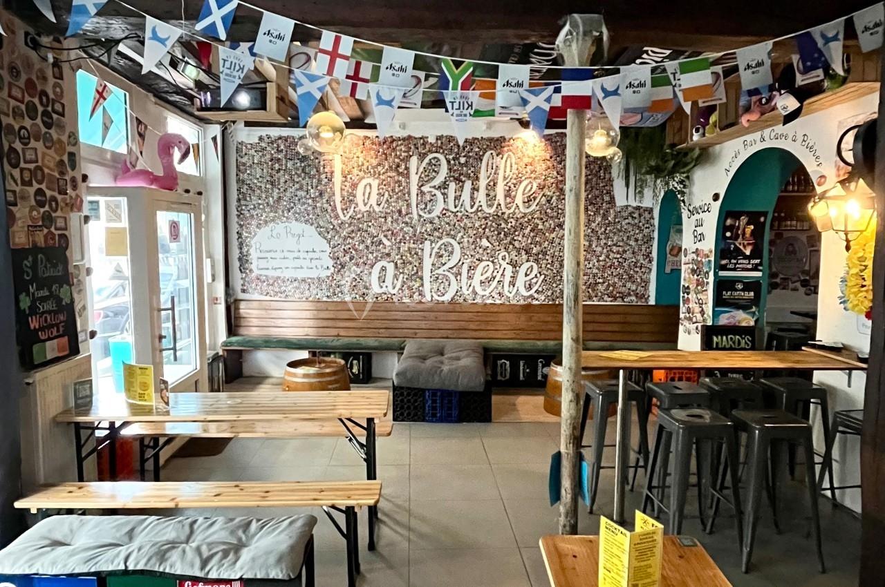 Intérieur d'un bar avec tables en bois, guirlandes de drapeaux et une enseigne murale ’La Bulle à Bière’.