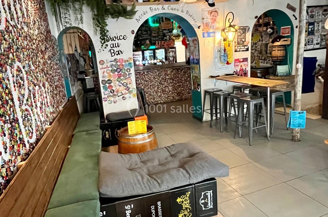 Intérieur d'un bar avec des tonneaux en tables, un banc, des drapeaux suspendus et un comptoir décoré au fond.