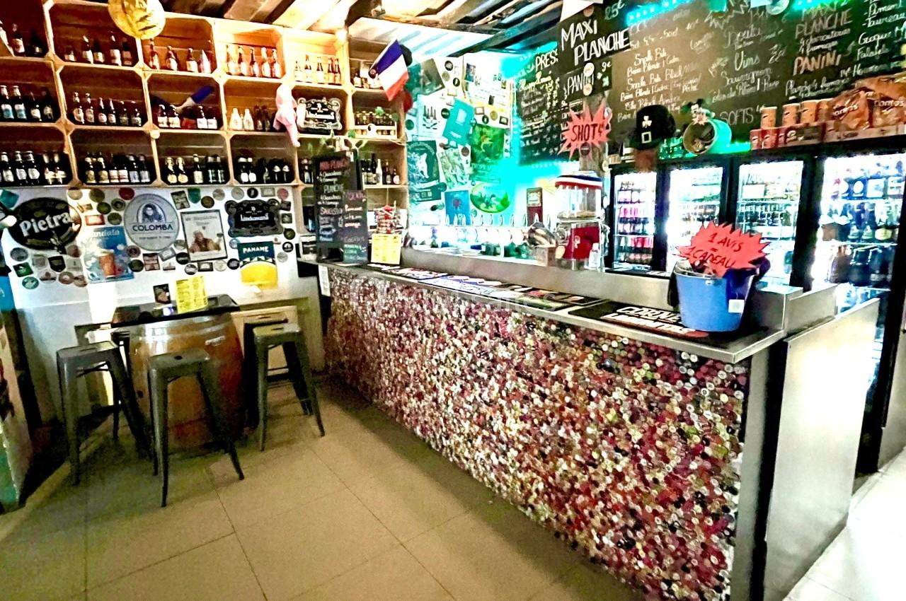 Intérieur d'un bar avec comptoir décoré de capsules, étagères remplies de bouteilles et drapeaux suspendus au plafond.