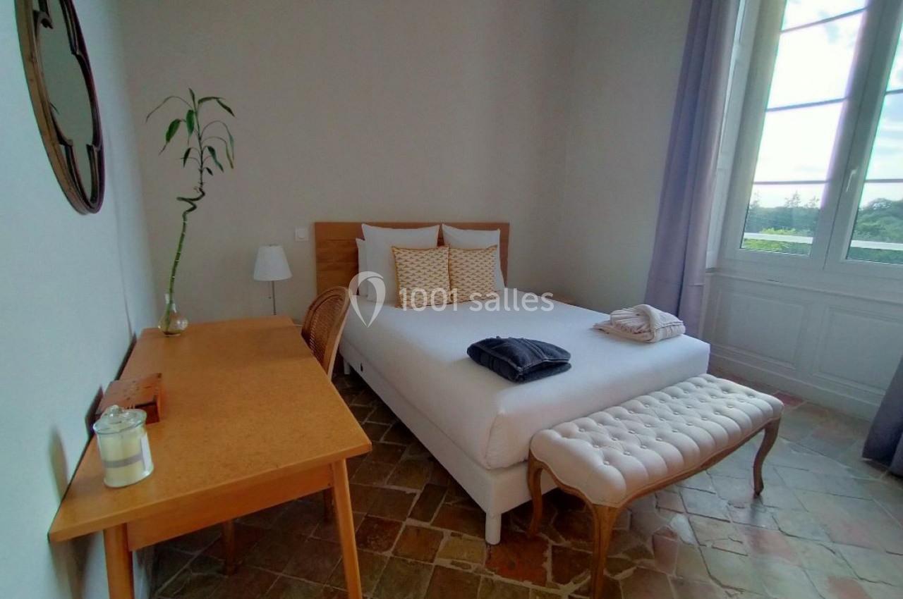 Chambre lumineuse avec lit double, bureau en bois, banc capitonné et grande fenêtre donnant sur un paysage extérieur.