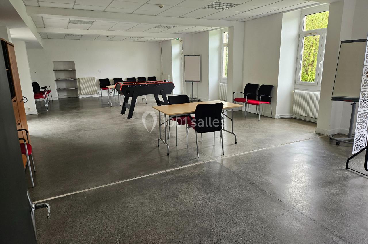 Salle lumineuse avec tables, chaises, baby-foot et tableaux blancs, sol en béton poli et grandes fenêtres.