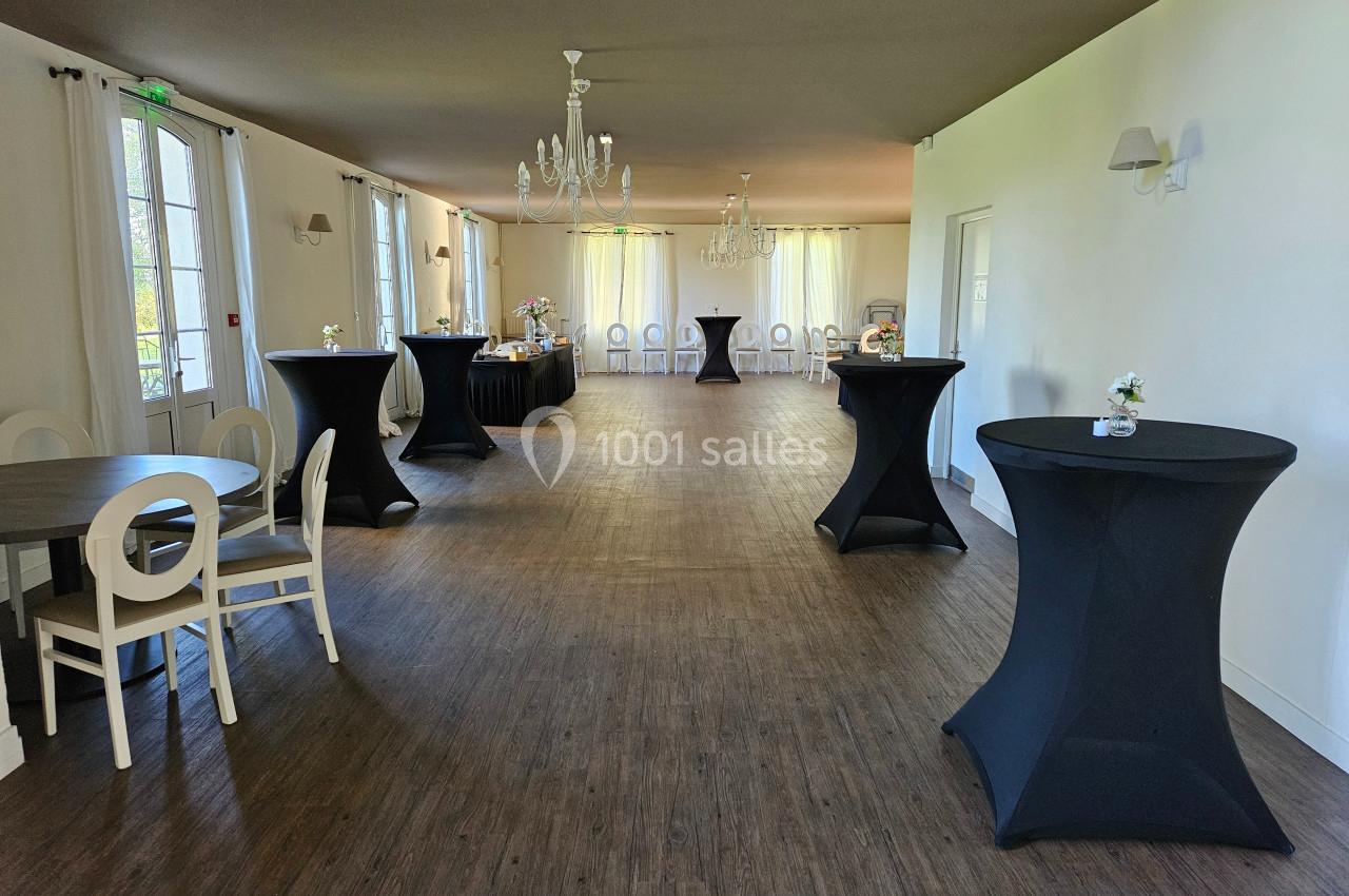 Salle lumineuse avec tables hautes noires, chaises blanches et décorations minimalistes, prête pour un événement.