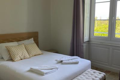 Chambre lumineuse avec un lit double, une grande fenêtre donnant sur un paysage verdoyant, et un fauteuil ancien.