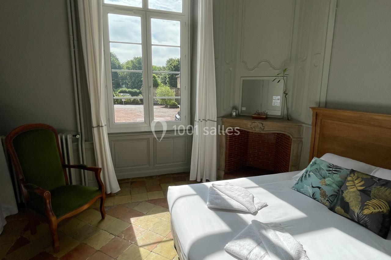 Chambre lumineuse avec un lit double, une grande fenêtre donnant sur un paysage verdoyant, et un fauteuil ancien.