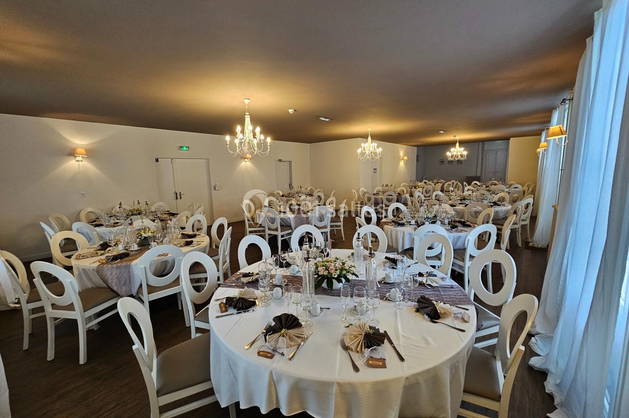 Salle de réception élégamment décorée avec des tables rondes dressées, chaises blanches et lumière naturelle.