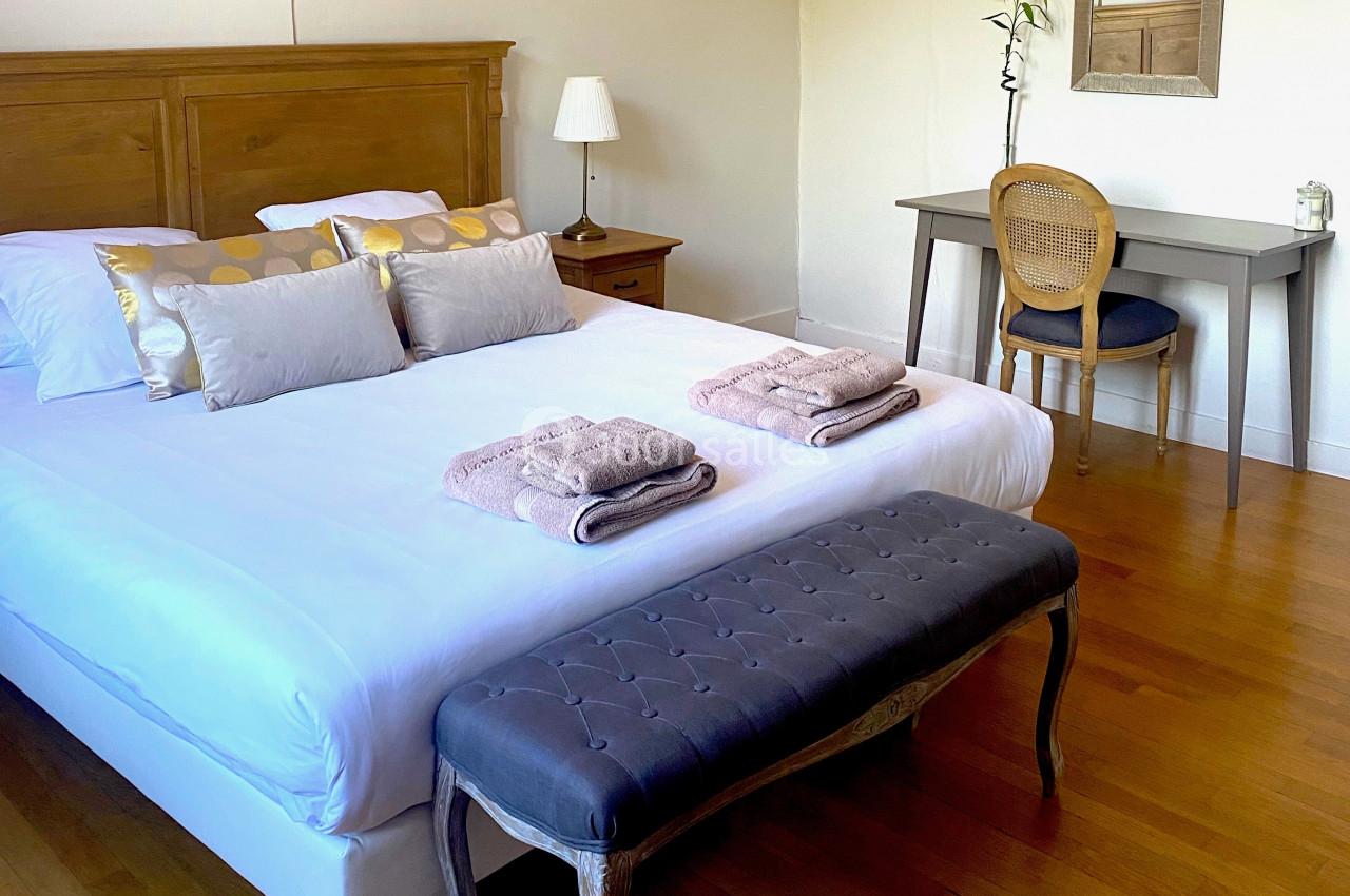 Chambre lumineuse avec lit double, banc capitonné, bureau en bois, lampe et miroir sur un parquet en bois clair.