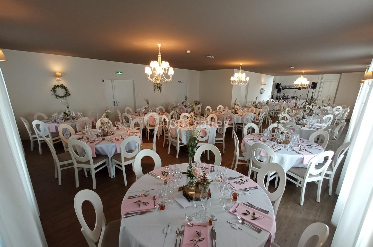 Salle de réception décorée avec des tables rondes, nappes blanches, serviettes roses et centres de table fleuris.