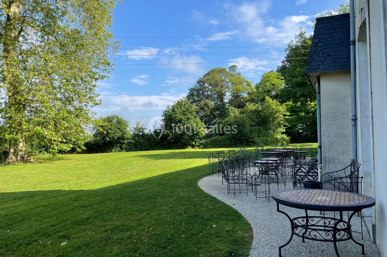 Terrasse avec tables et chaises en fer forgé, bordée d'une pelouse et d'arbres sous un ciel dégagé.