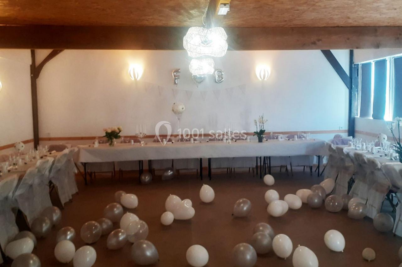 Salle décorée pour une réception avec longues tables dressées, ballons blancs et argentés dispersés au sol.