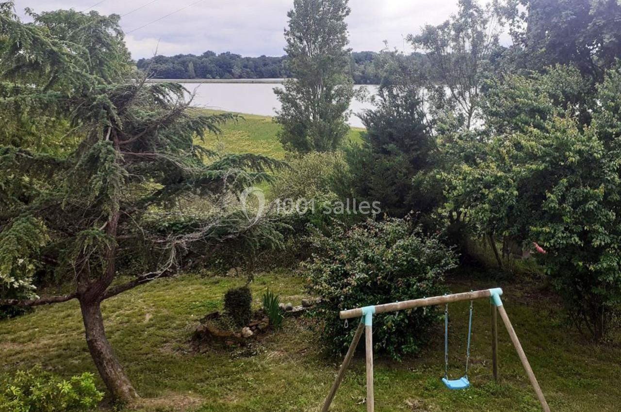 Vue d'un jardin verdoyant avec une balançoire en bois, des arbres et un lac en arrière-plan.