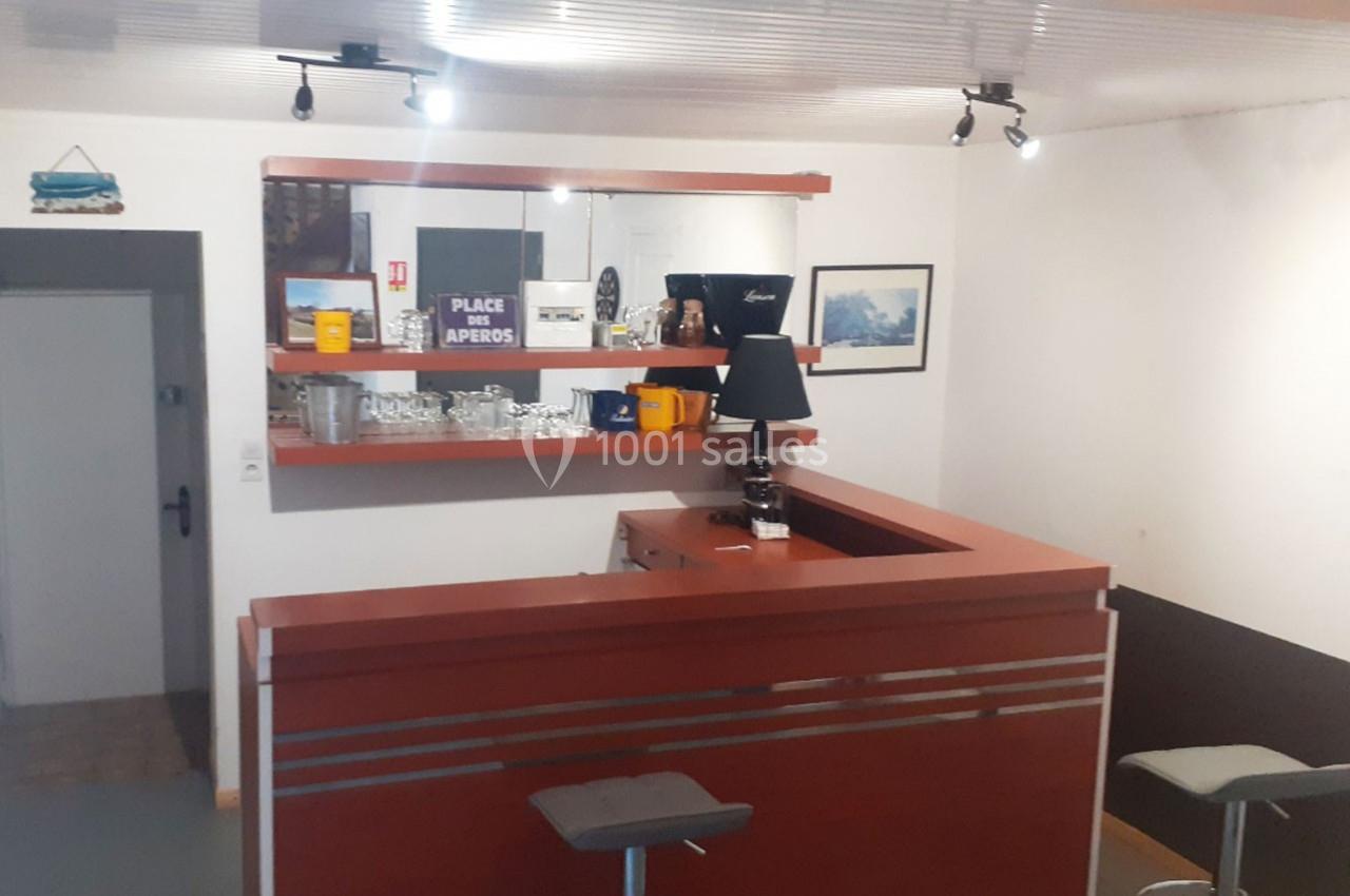 Comptoir de bar en bois avec tabourets, étagères contenant verres et objets décoratifs, éclairage au plafond.