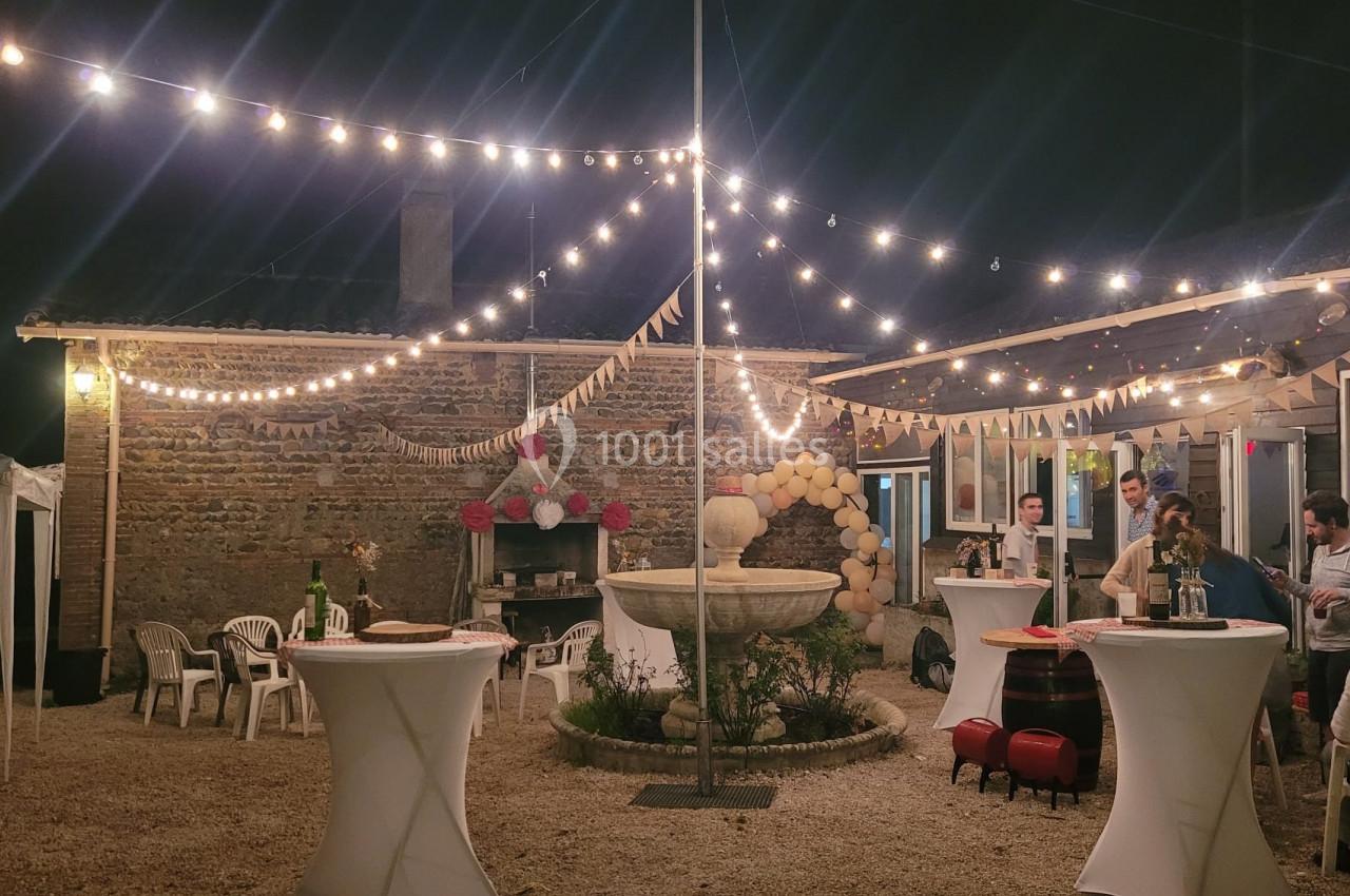 Cour extérieure éclairée par des guirlandes lumineuses, décorée pour une fête avec tables hautes et fontaine centrale.