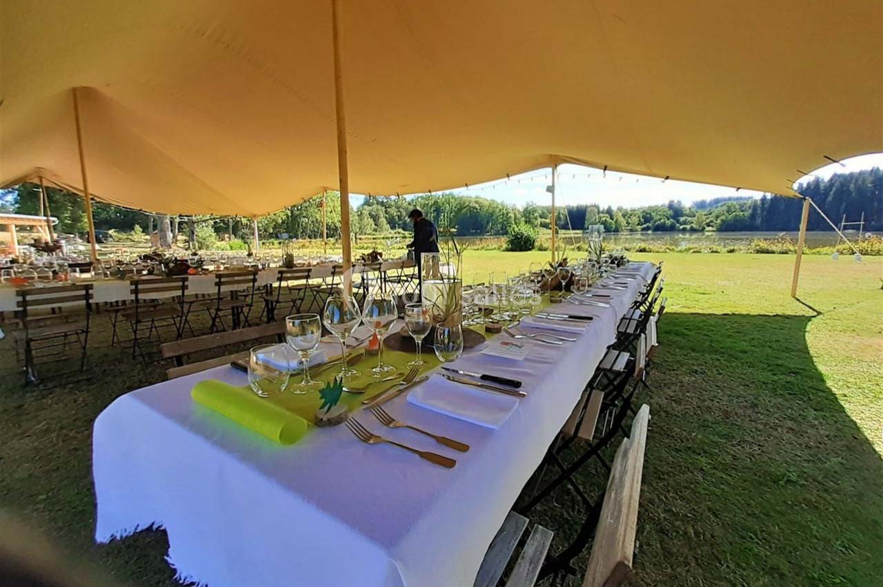 Table dressée sous une tente, avec nappes blanches, couverts et décorations, dans un cadre champêtre ensoleillé.