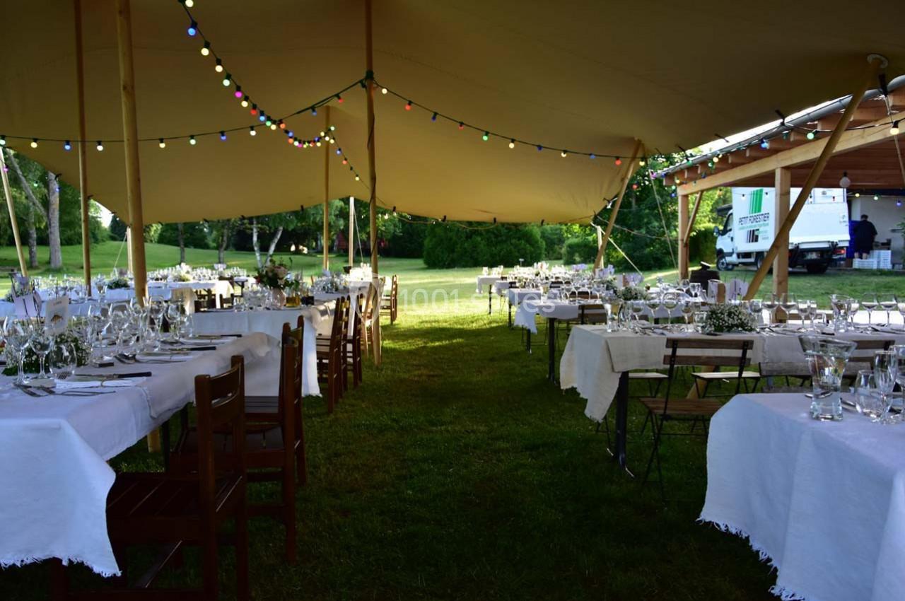 Tables dressées sous une tente éclairée par des guirlandes lumineuses, installée dans un espace extérieur verdoyant.