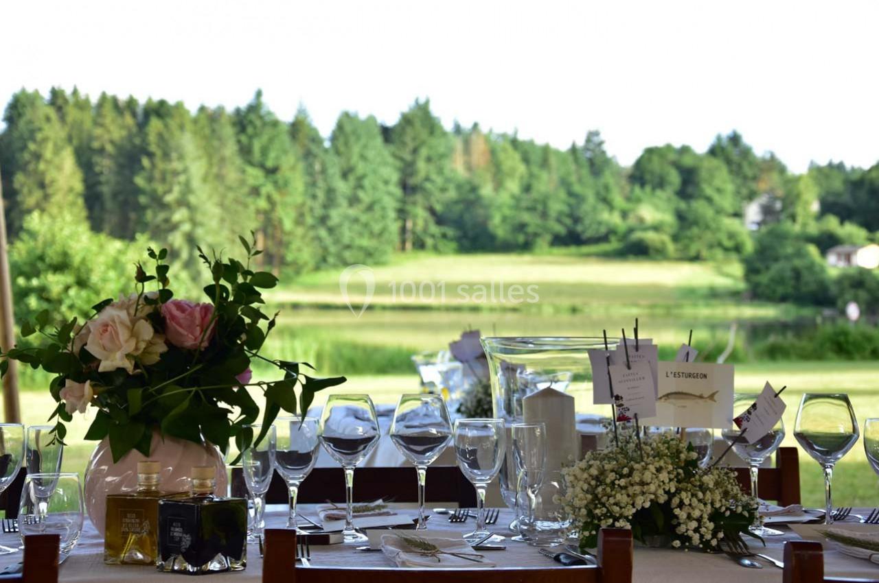 Table élégamment dressée avec des verres, des fleurs et des menus, offrant une vue sur un paysage verdoyant.
