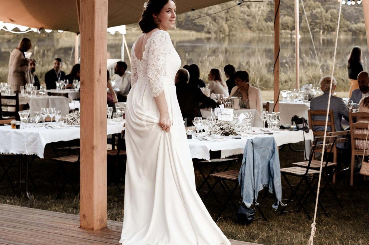 Une femme en robe de mariée blanche se tient sous une tente lors d'une réception en plein air près d'un lac.