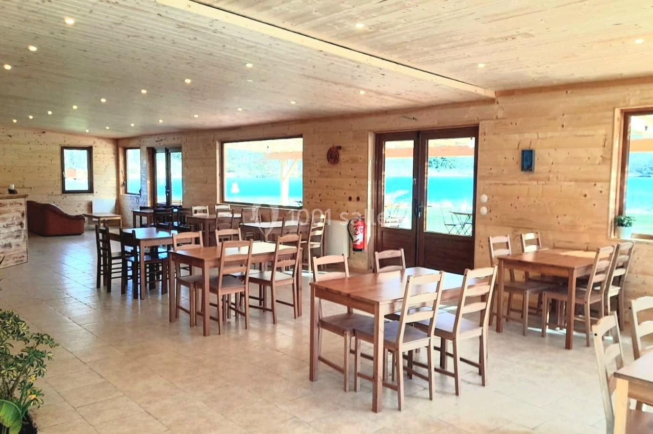 Salle lumineuse en bois avec tables et chaises, grandes fenêtres donnant sur un extérieur ensoleillé.