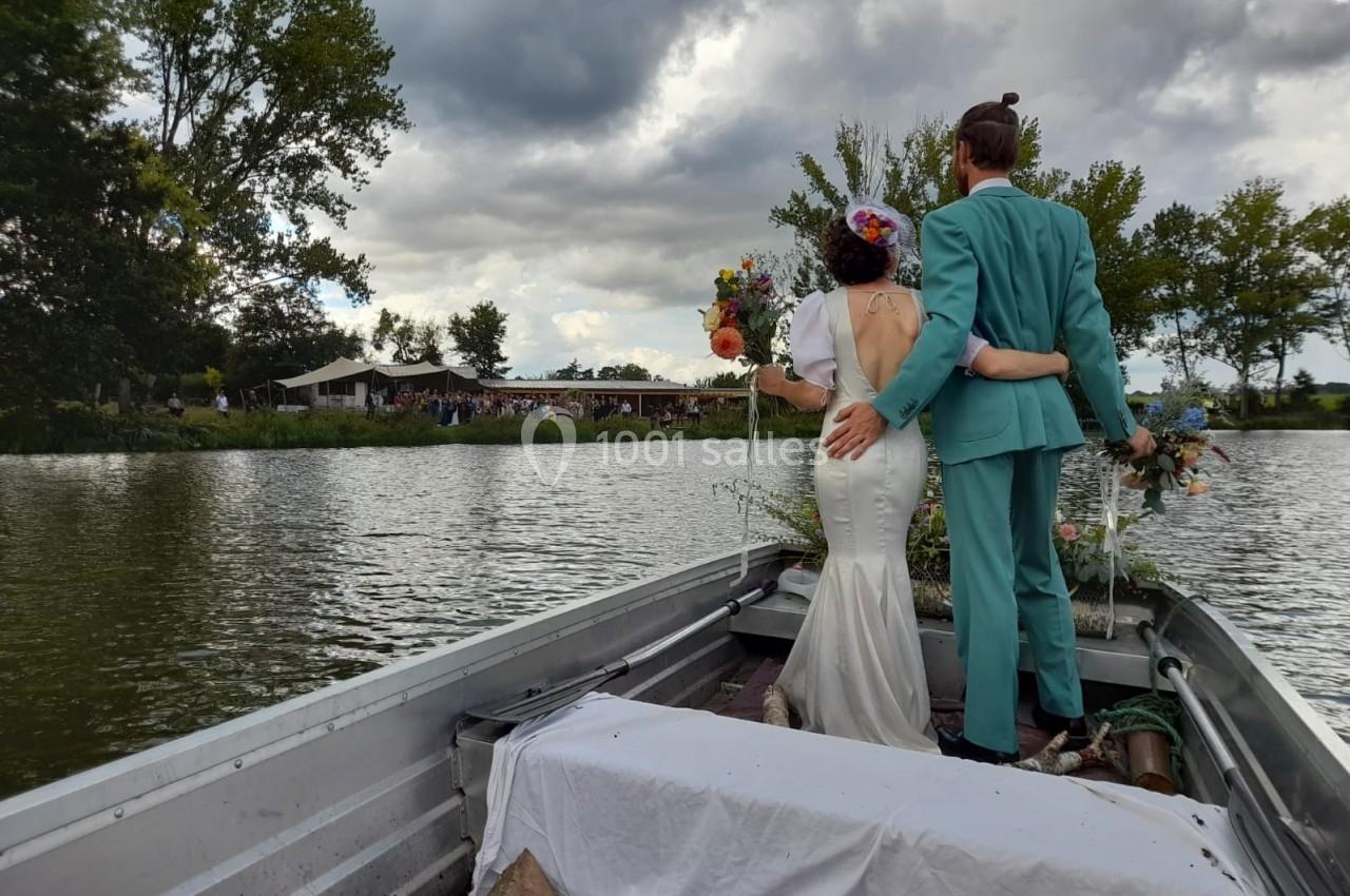 Un couple en tenue de mariage se tient debout à l'arrière d'une barque sur un lac, entouré de végétation.