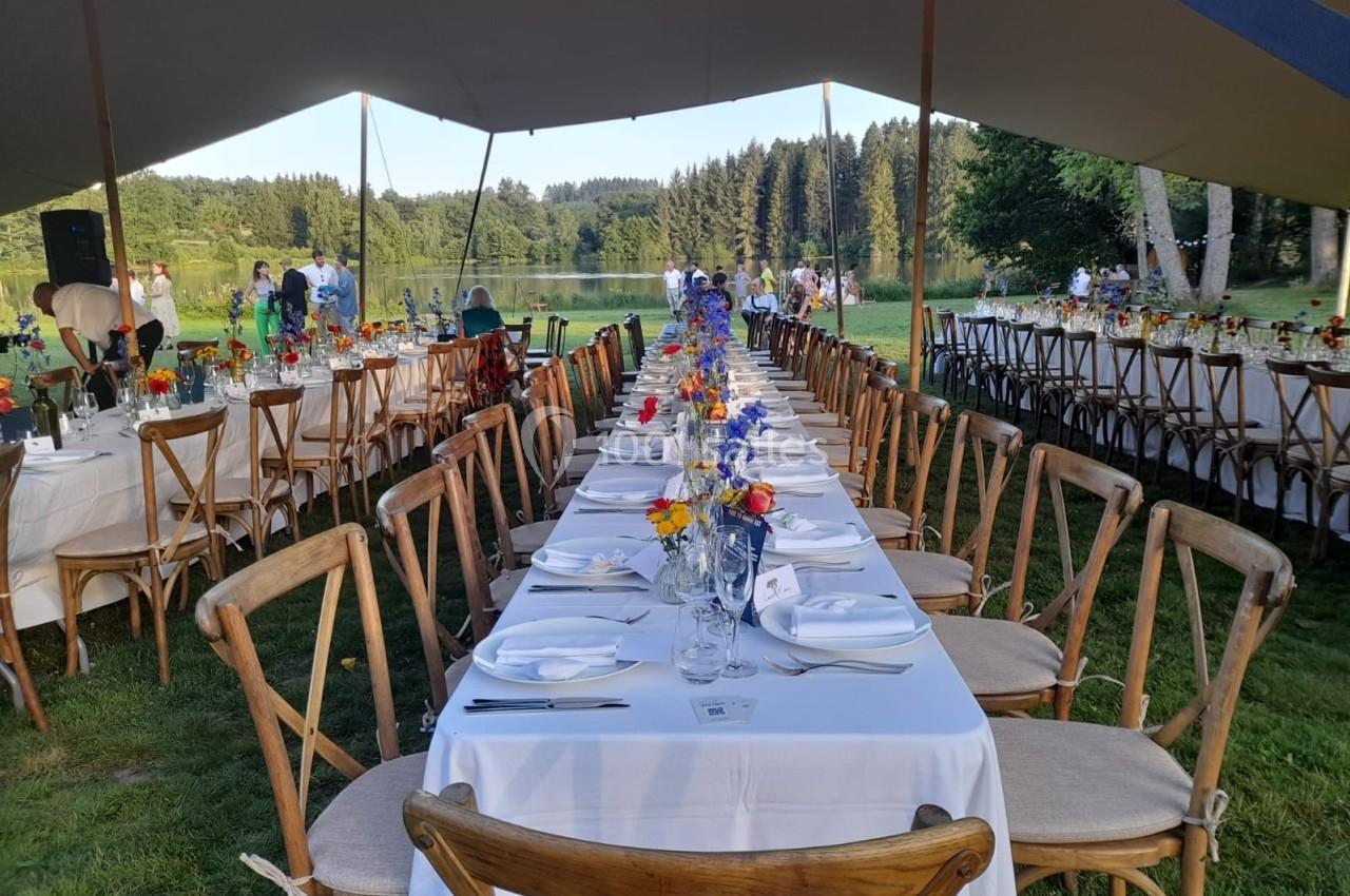 Tables dressées en extérieur sous une tente, avec nappes blanches, chaises en bois et bouquets de fleurs colorées.