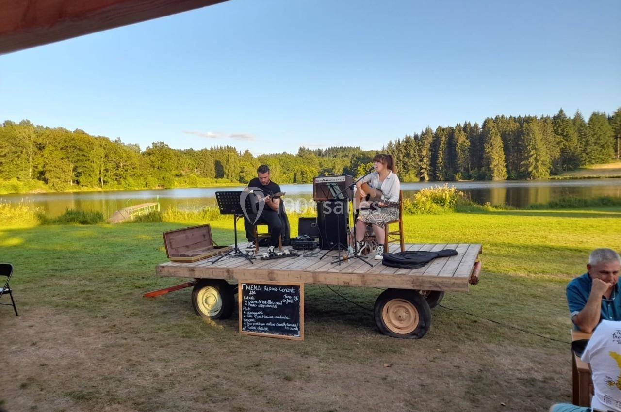 Deux musiciens jouent sur une petite scène en plein air près d'un lac entouré de forêt.