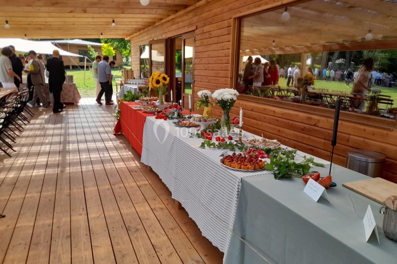 Buffet varié dressé sur des tables en extérieur sous une terrasse en bois, avec des invités en arrière-plan.