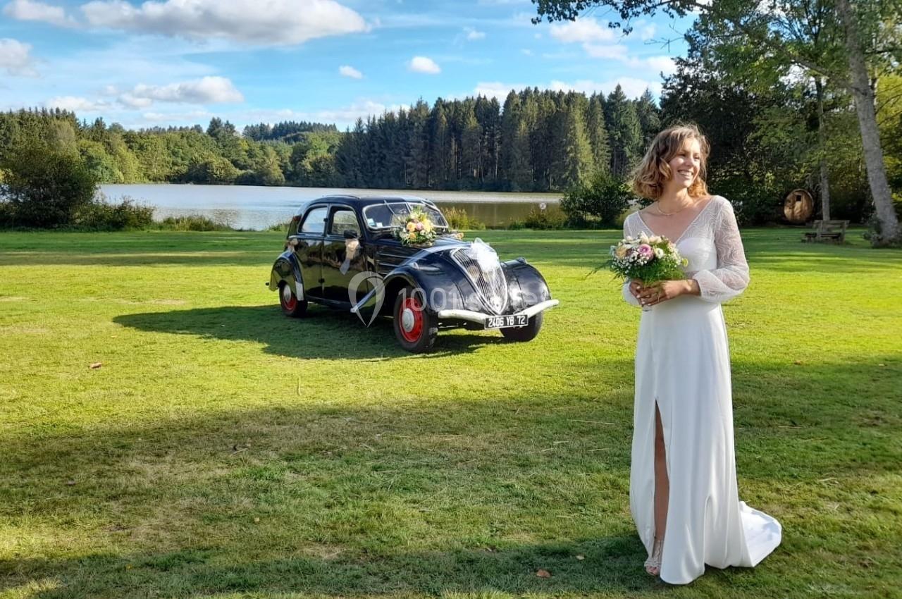 Une femme en robe de mariée tient un bouquet sur une pelouse, avec une voiture ancienne et un lac en arrière-plan.