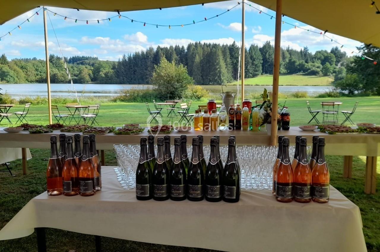 Tables dressées en extérieur avec bouteilles de vin, verres, boissons et collations, près d'un lac entouré de forêt.