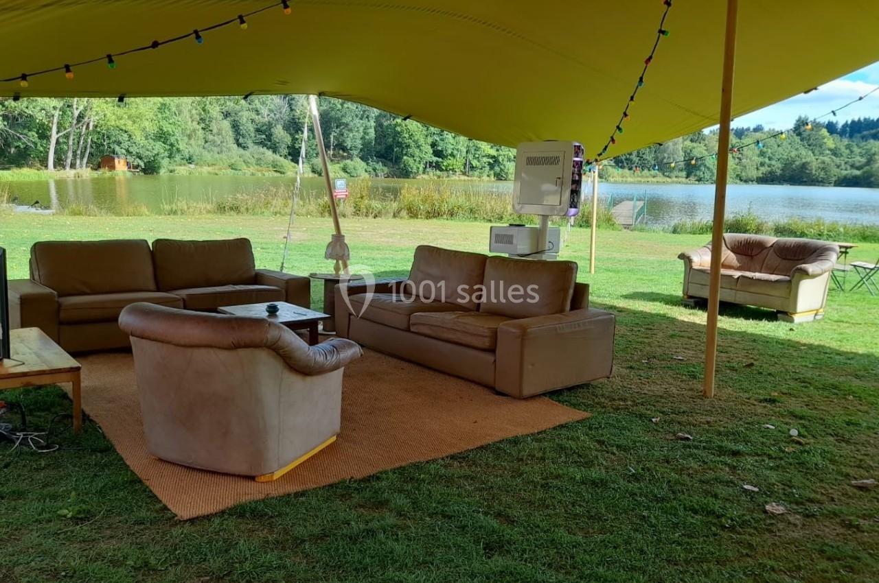 Salon extérieur sous une tente jaune, avec canapés et fauteuils disposés sur un tapis, près d'un lac entouré de verdure.