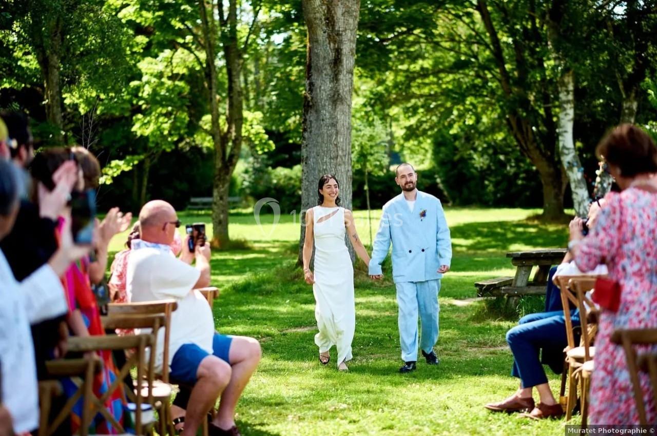 Un couple marche main dans la main dans une allée extérieure entourée d'invités assis, sous des arbres verdoyants.