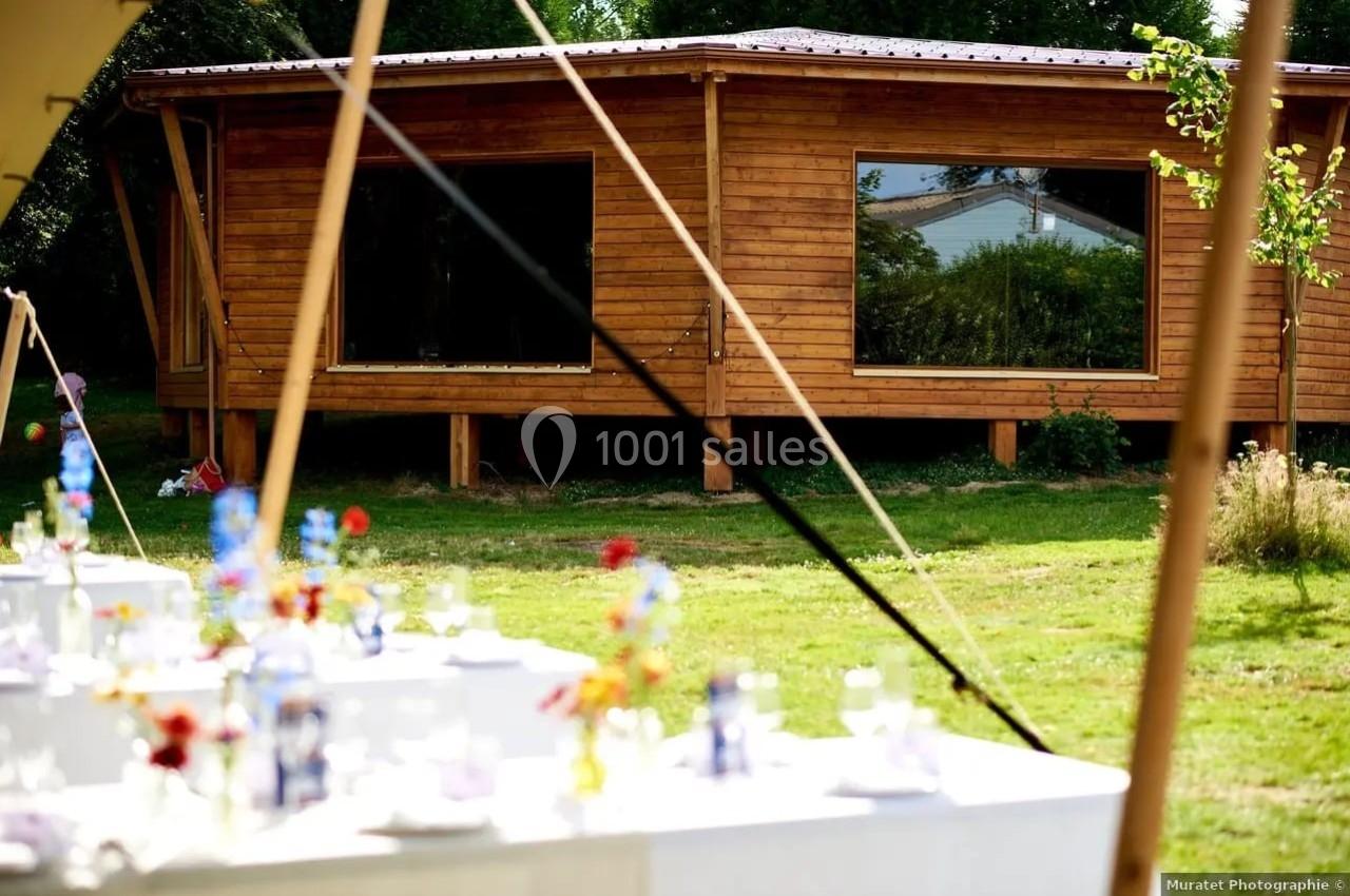 Salle en bois avec de grandes fenêtres, entourée de verdure, avec des tables décorées pour un événement au premier plan.
