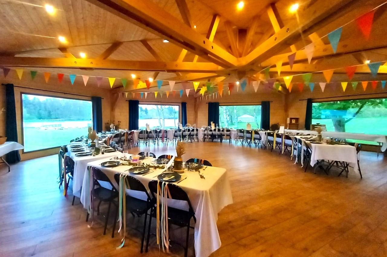 Salle de réception en bois décorée de guirlandes colorées, avec des tables dressées pour un événement festif.