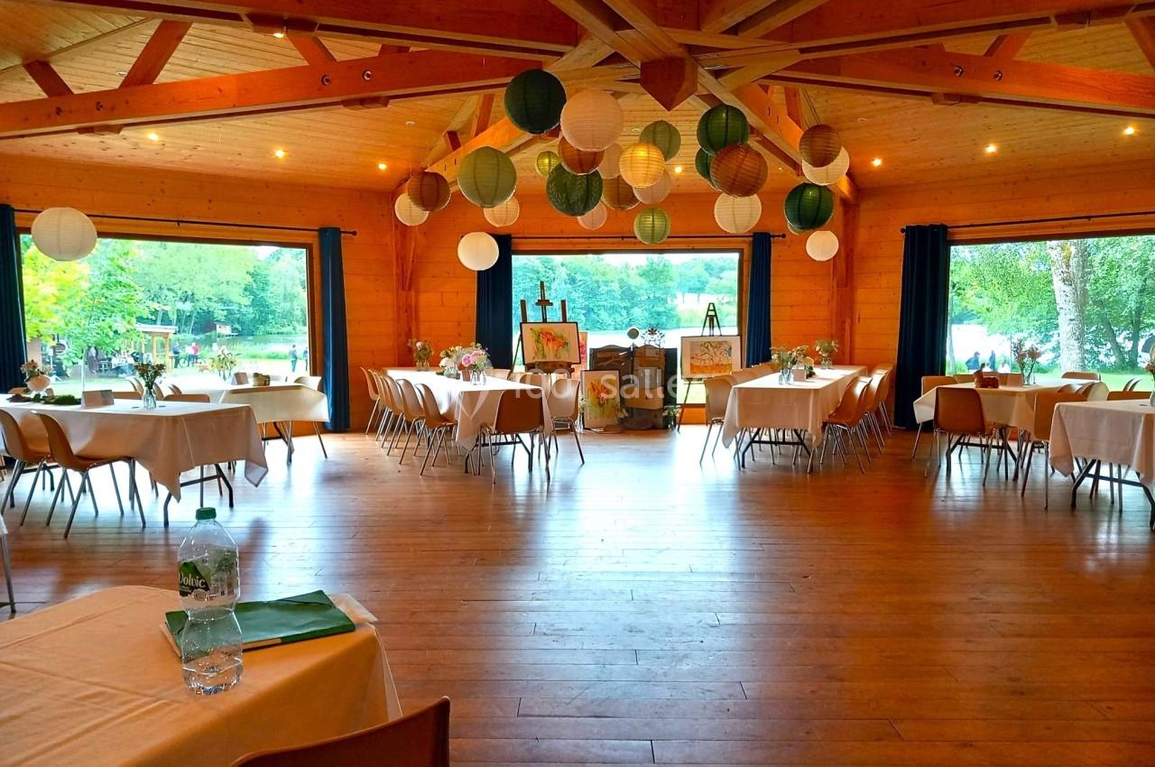Salle en bois avec tables dressées, lanternes suspendues et vue sur un espace extérieur verdoyant à travers de grandes…