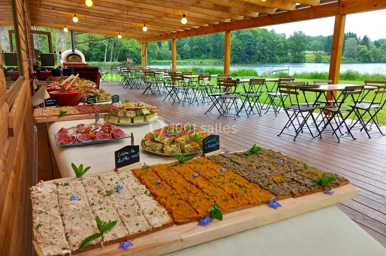 Buffet en plein air avec tartines variées et tables installées sous une pergola en bois près d'un lac.