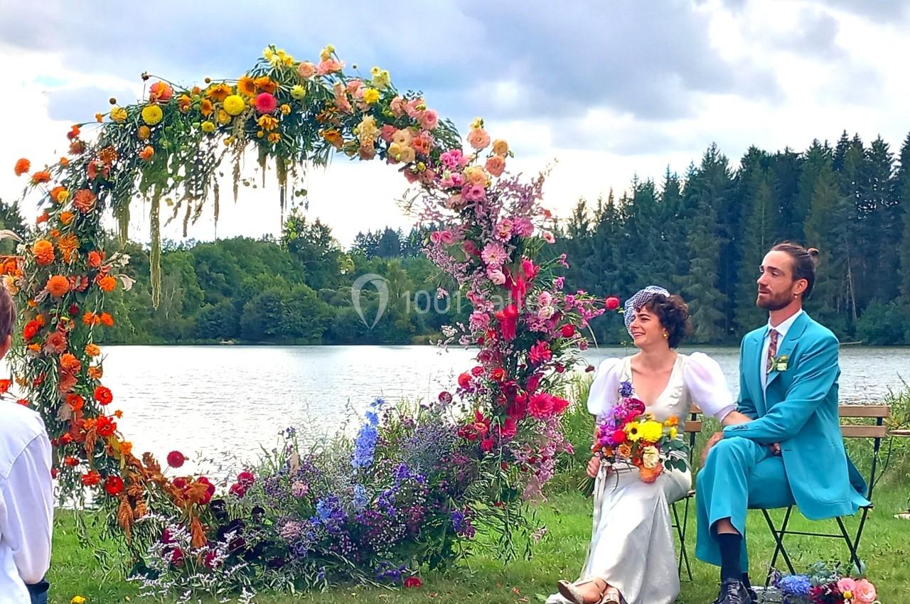 Un couple assis près d'une arche florale colorée au bord d'un lac, entouré de nature et d'un ciel nuageux.