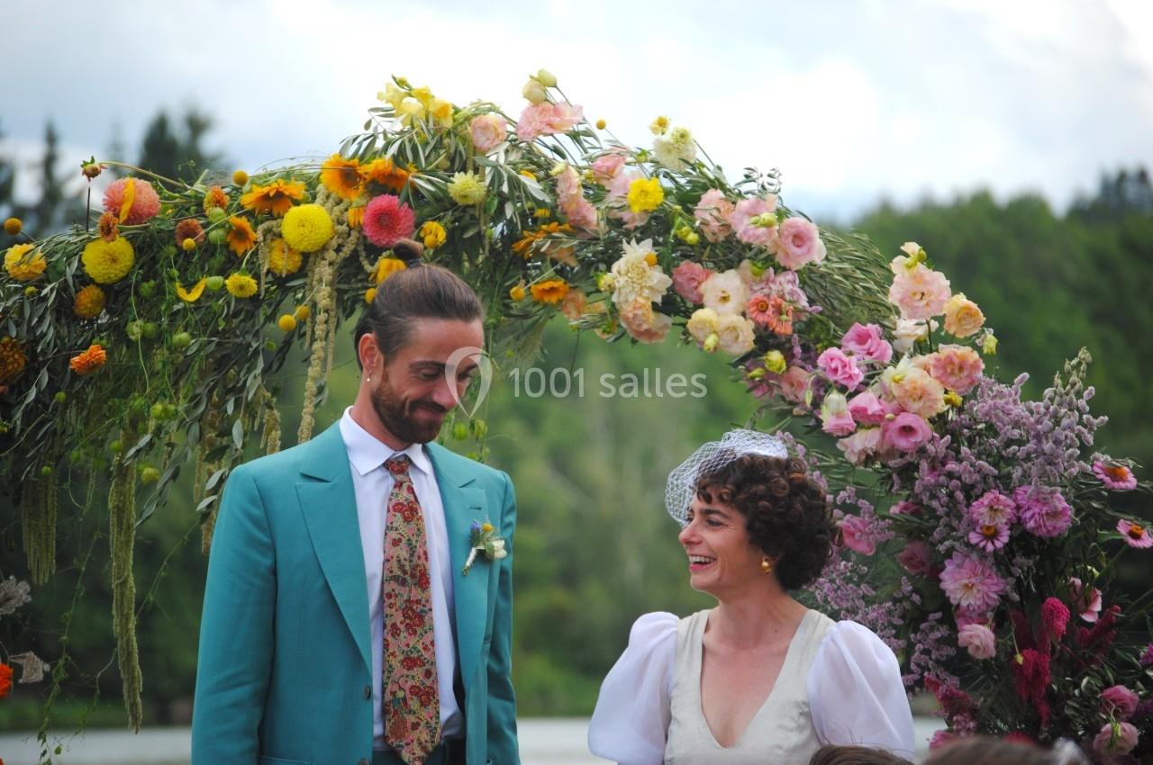 Un couple souriant sous une arche florale colorée lors d'une cérémonie en plein air, avec un paysage naturel en arrière-plan.