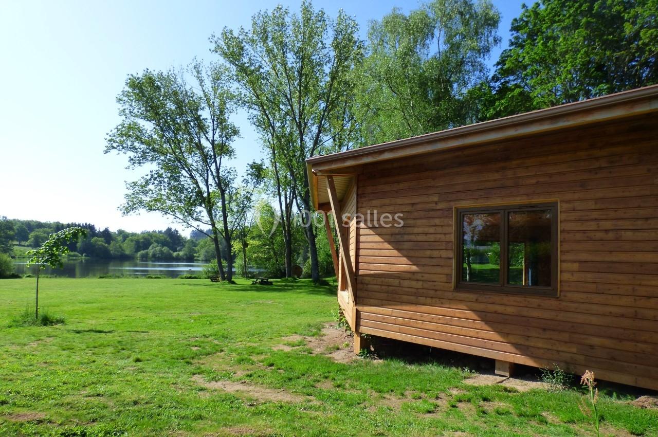 Chalet en bois au bord d'un lac entouré de pelouse et d'arbres sous un ciel dégagé.