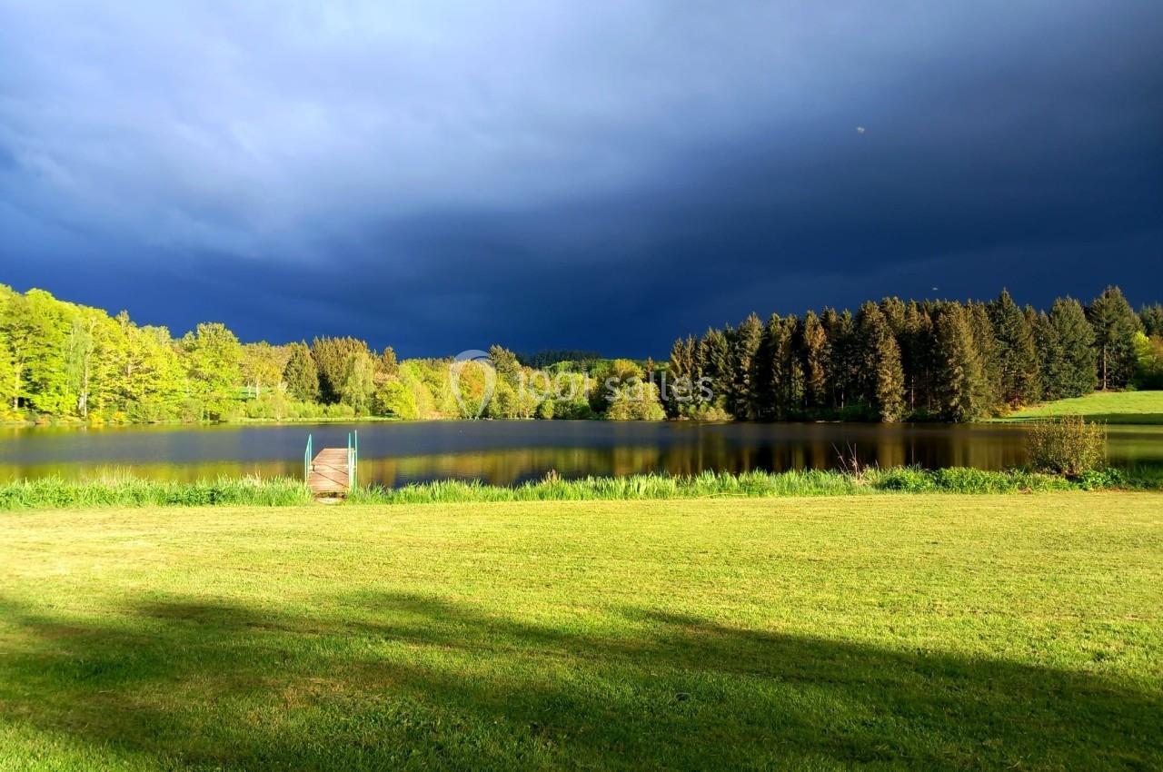 Paysage avec un lac entouré de forêts et d'herbe verte, sous un ciel sombre et nuageux.