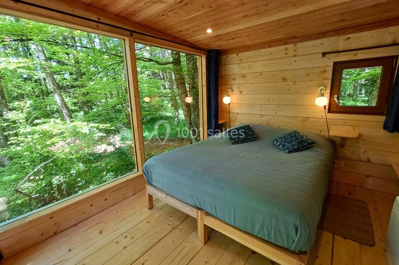 Chambre en bois avec un grand lit, éclairée par une baie vitrée donnant sur une forêt verdoyante.