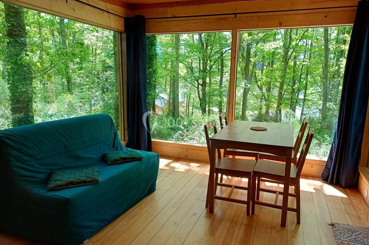 Salon lumineux avec canapé, table en bois et grandes baies vitrées offrant une vue sur une forêt verdoyante.
