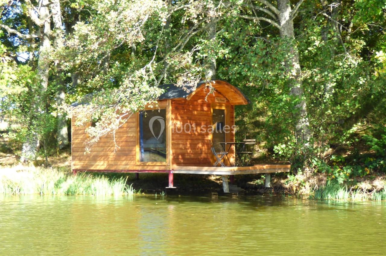 Cabane en bois sur pilotis au bord d'un lac, entourée d'arbres et baignée par la lumière du soleil.