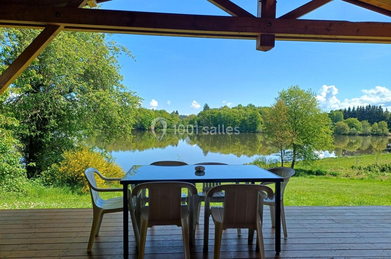Vue sur un lac entouré de verdure depuis une terrasse en bois avec une table et des chaises en plastique.
