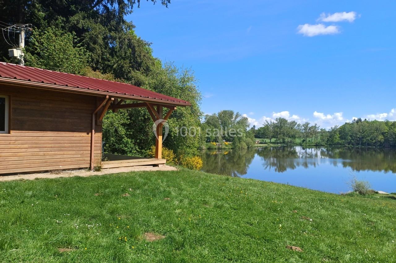 Cabane en bois avec toit rouge près d'un lac entouré de verdure sous un ciel bleu.