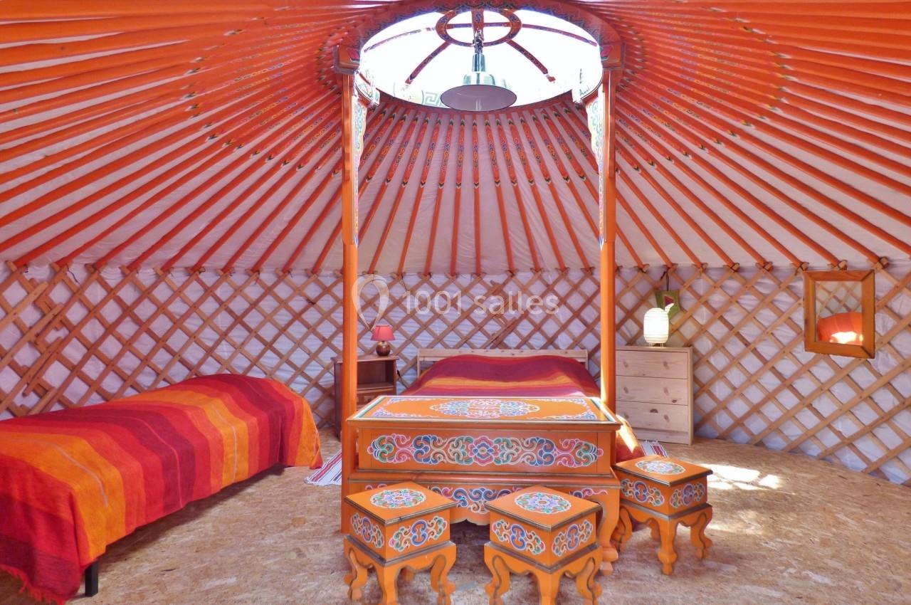Intérieur d'une yourte avec un lit, des tabourets décorés, une table basse et des murs en bois entrecroisés.