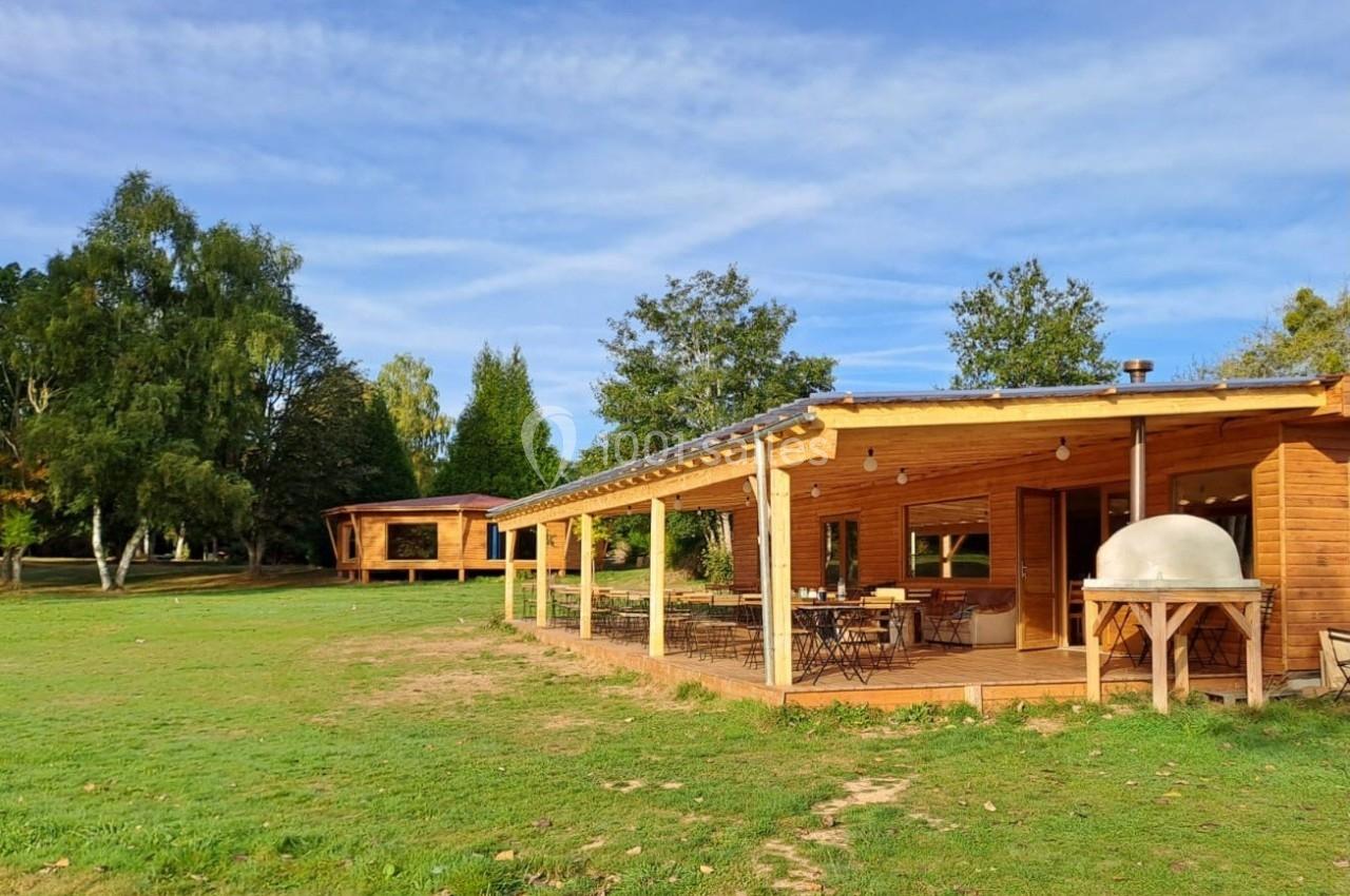 Terrasse en bois d'un bâtiment avec tables et chaises, entourée d'un grand espace vert et d'arbres sous un ciel bleu.