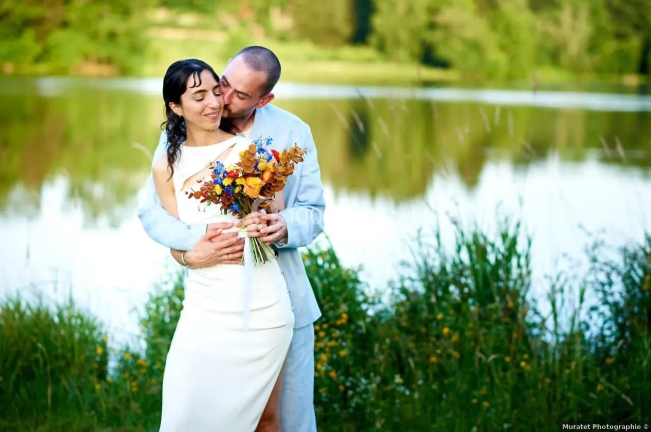 Un couple en tenue de mariage pose près d'un lac, entouré de verdure, avec un bouquet coloré.