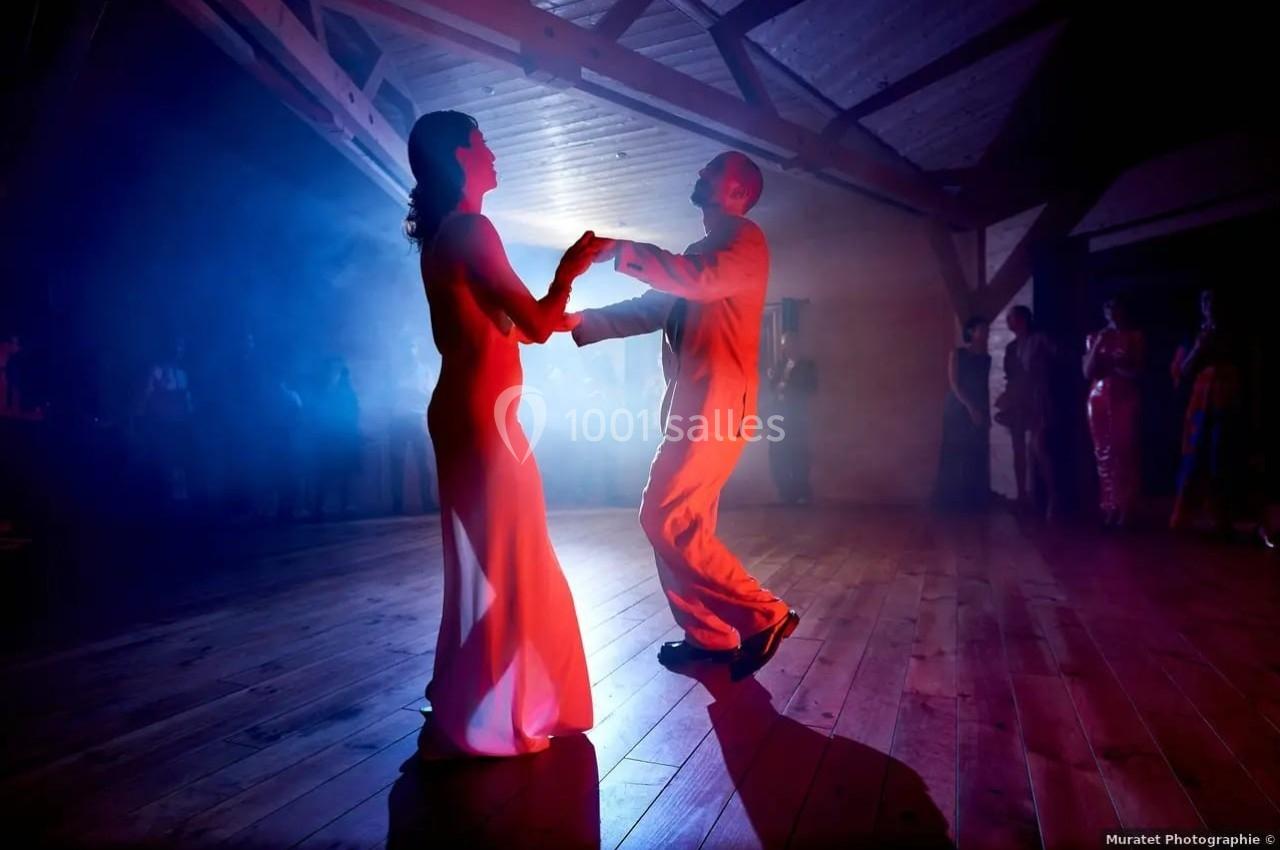 Un couple danse sous des lumières colorées dans une salle au parquet en bois, entouré de quelques spectateurs.
